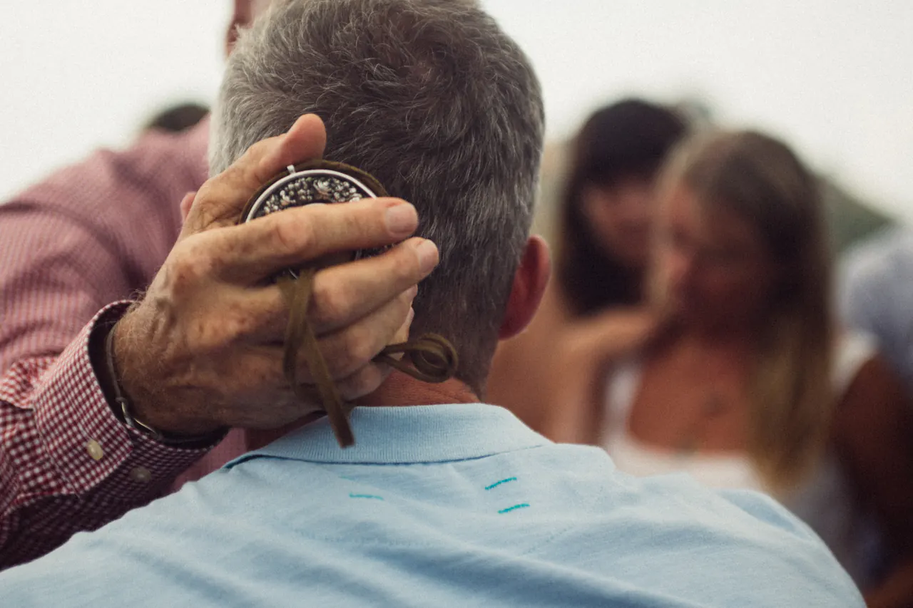 Lama Ole Nydahl presses a round silver reliquary on a leather strap to the back of a man's head in a blessing, a crowd blurred behind