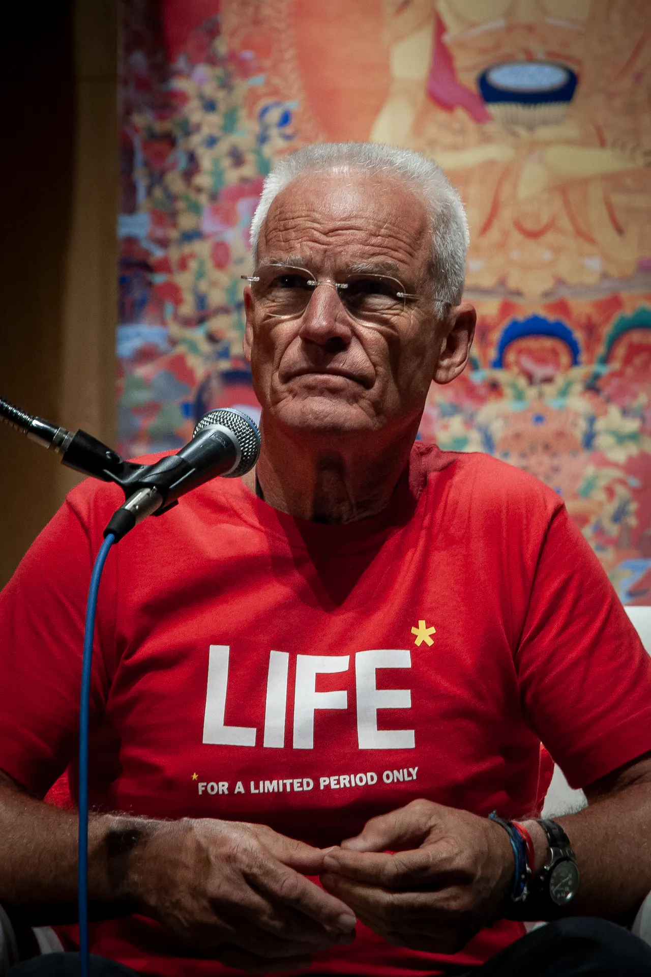 Lama Ole Nydahl in a red LIFE t-shirt sits at a microphone, a painted thangka behind him