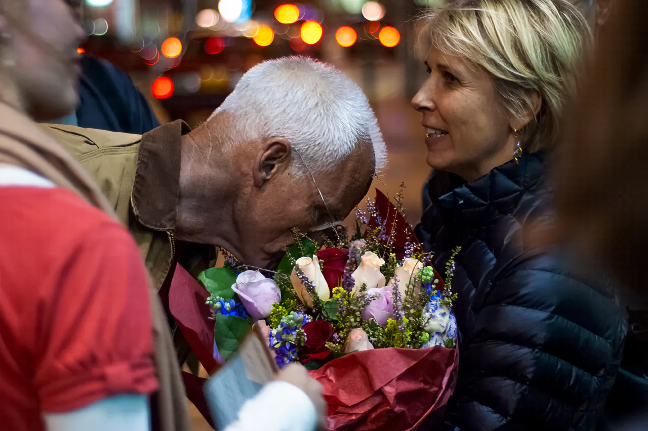 Lama Ole Nydahl bows his head towards a woman holding a bouquet, street lights blurred behind them