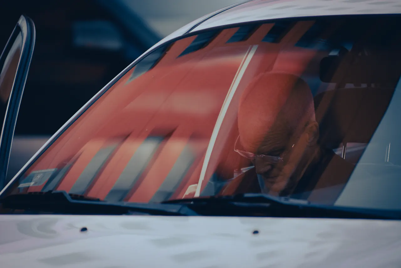Seen through a windscreen, Lama Ole Nydahl sits in a car in a red shirt