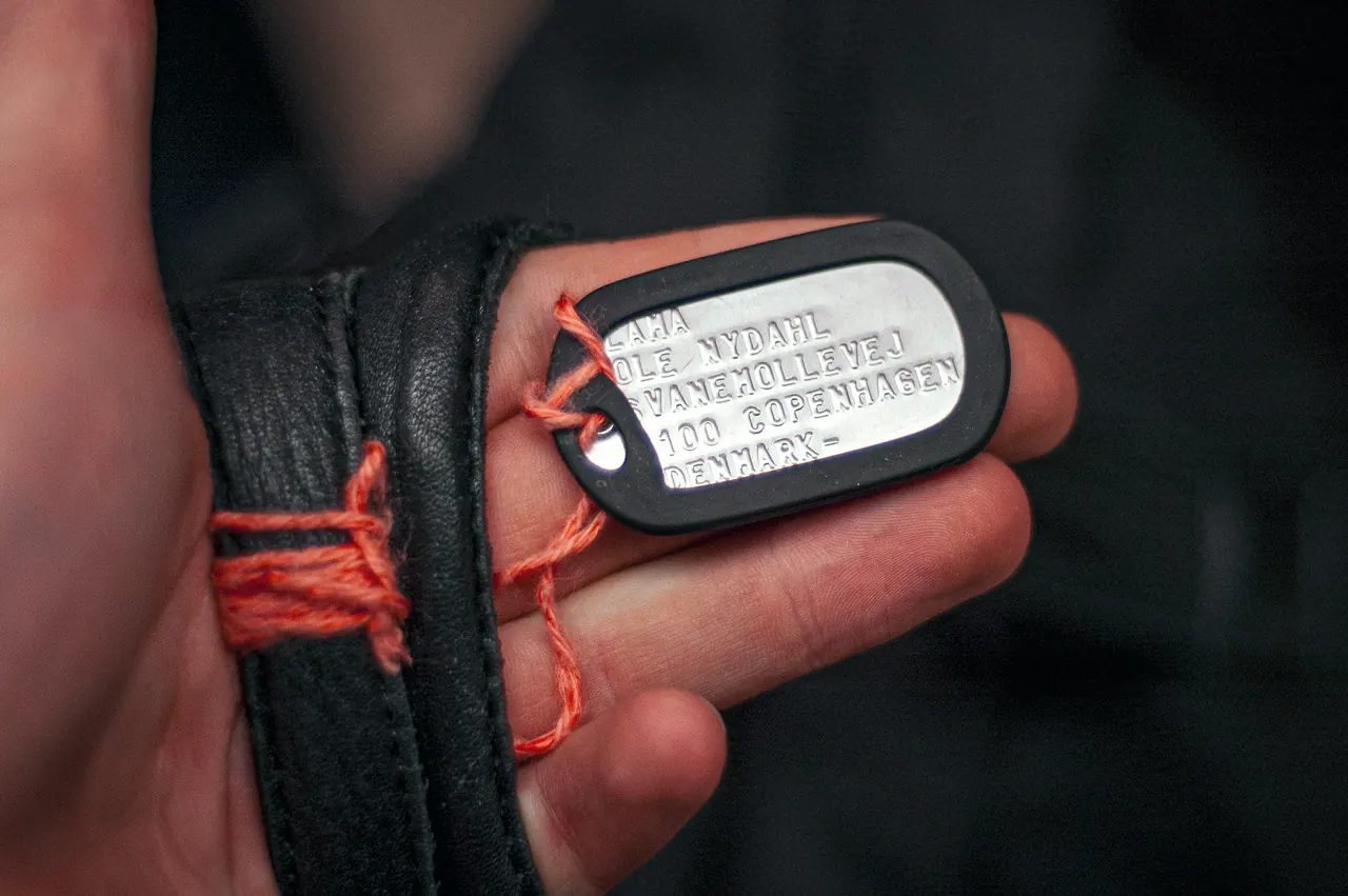 A hand holds up Lama Ole Nydahl's dog tag, engraved with his name and a Copenhagen address