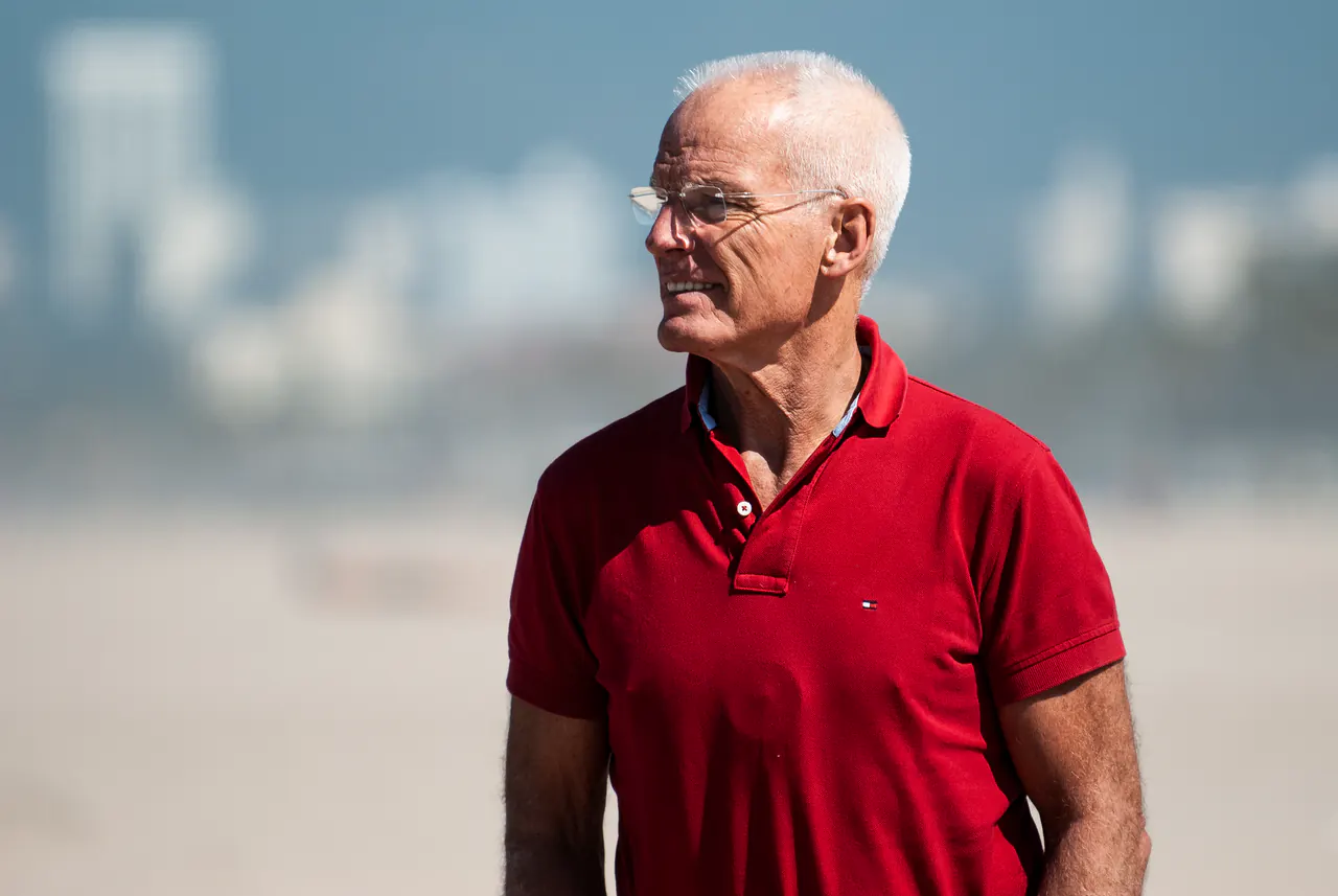 Lama Ole Nydahl in a red polo shirt on a wide beach, a hazy city skyline behind him