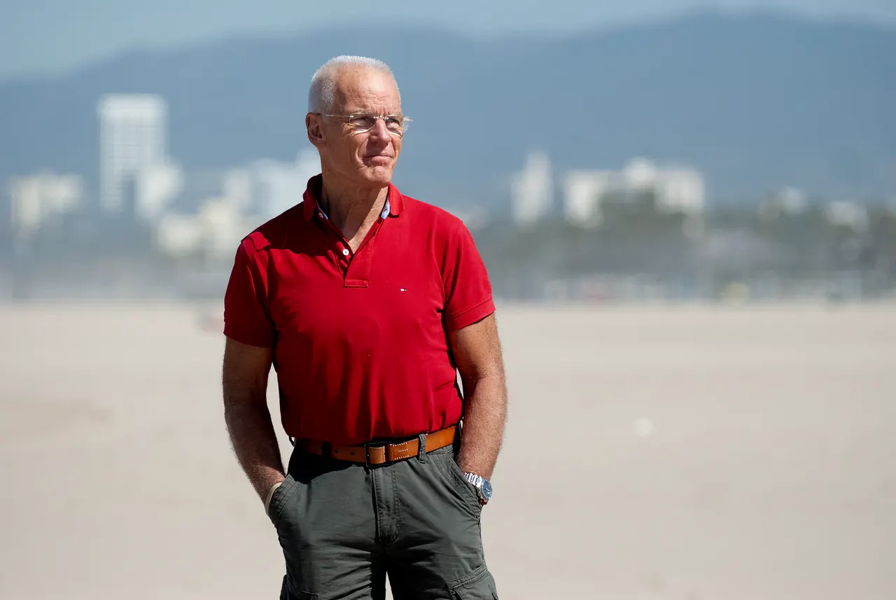 Lama Ole Nydahl stands with his hands in his pockets on the sand, the skyline blurred behind