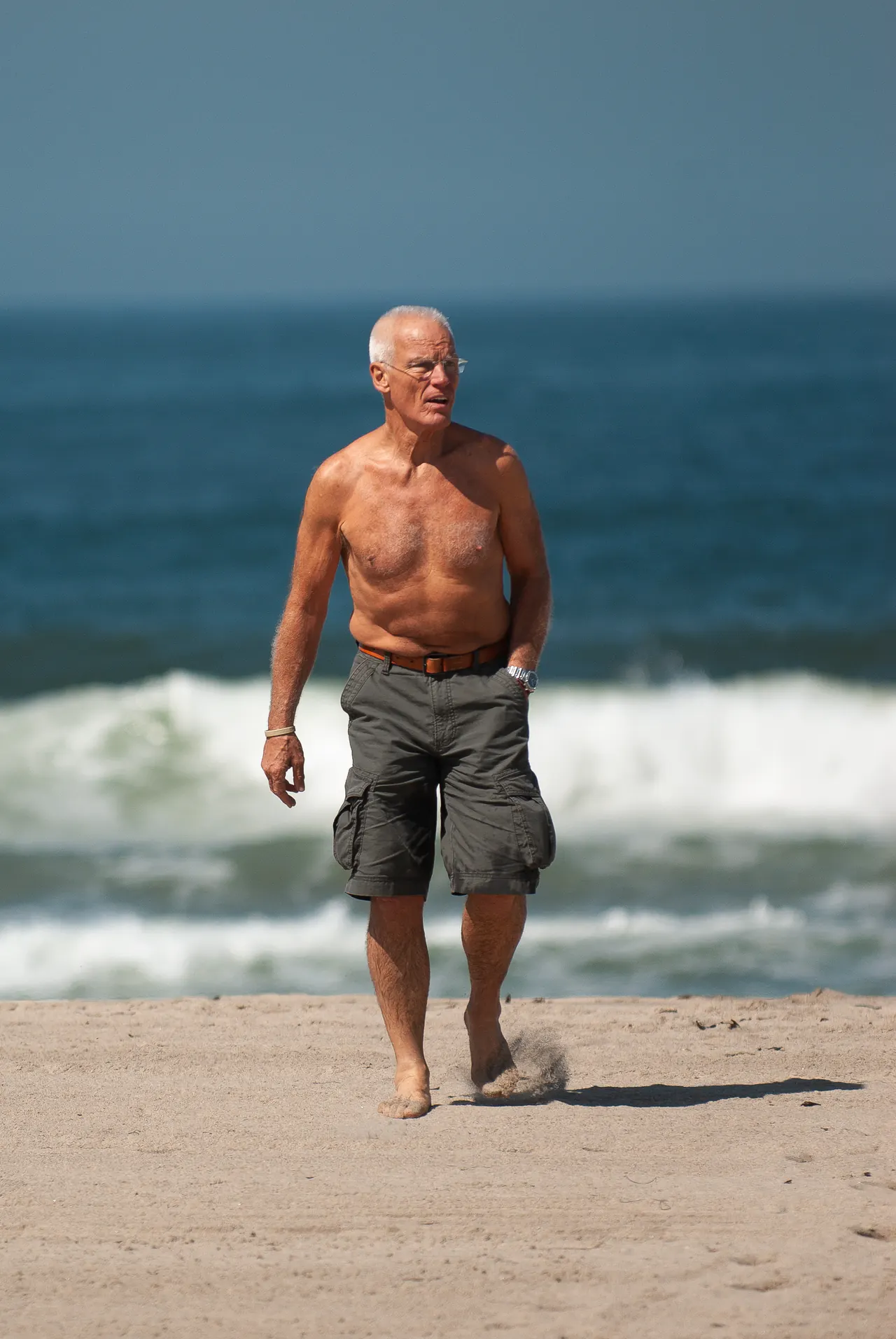 Bare-chested in cargo shorts, Lama Ole Nydahl walks barefoot up the beach from the surf