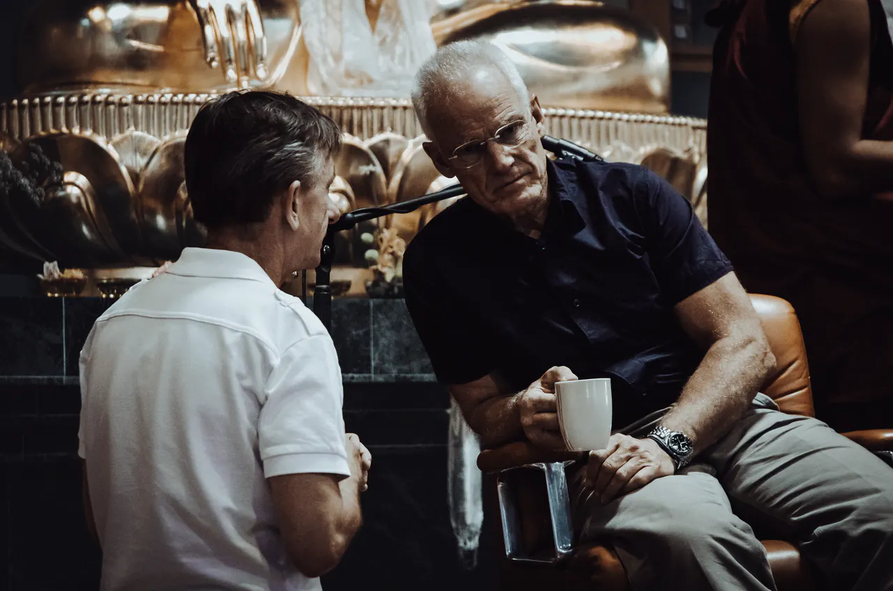 Lama Ole Nydahl sits with a mug in front of a large golden Buddha statue, talking with a man beside him