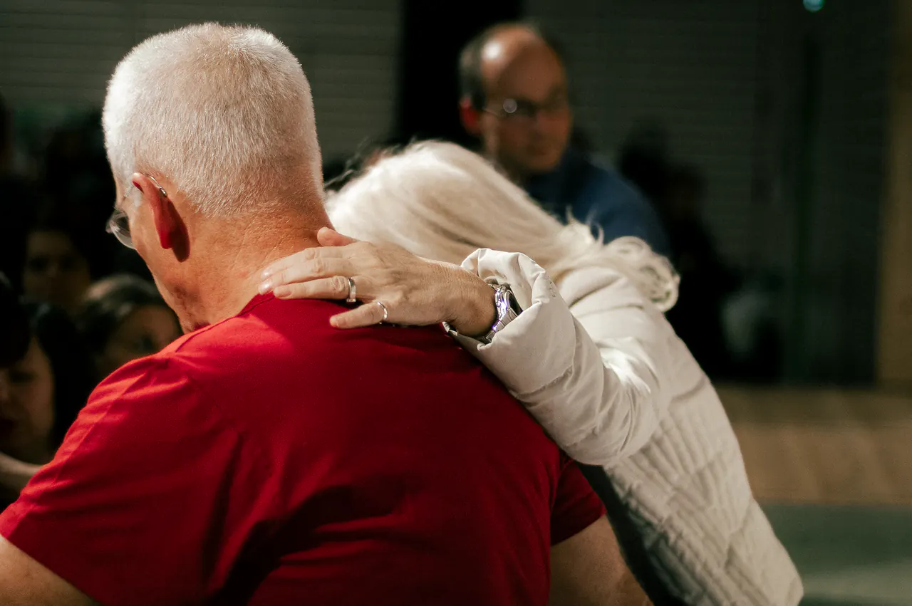 Seen from behind, Lama Ole Nydahl in a red t-shirt embraces a blonde woman in front of a seated audience