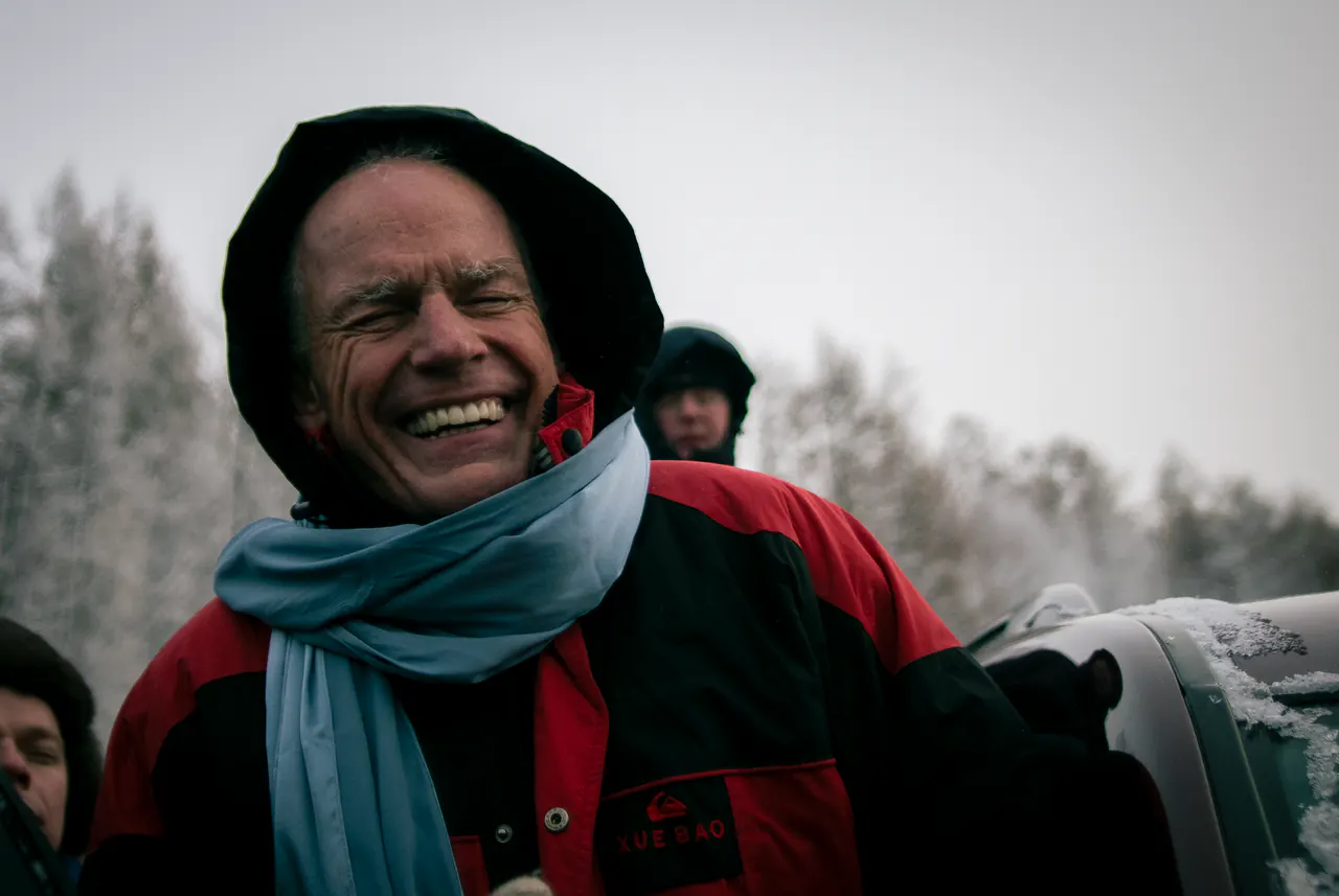 Lama Ole Nydahl laughs in a hooded jacket and scarf, snow-covered trees behind him
