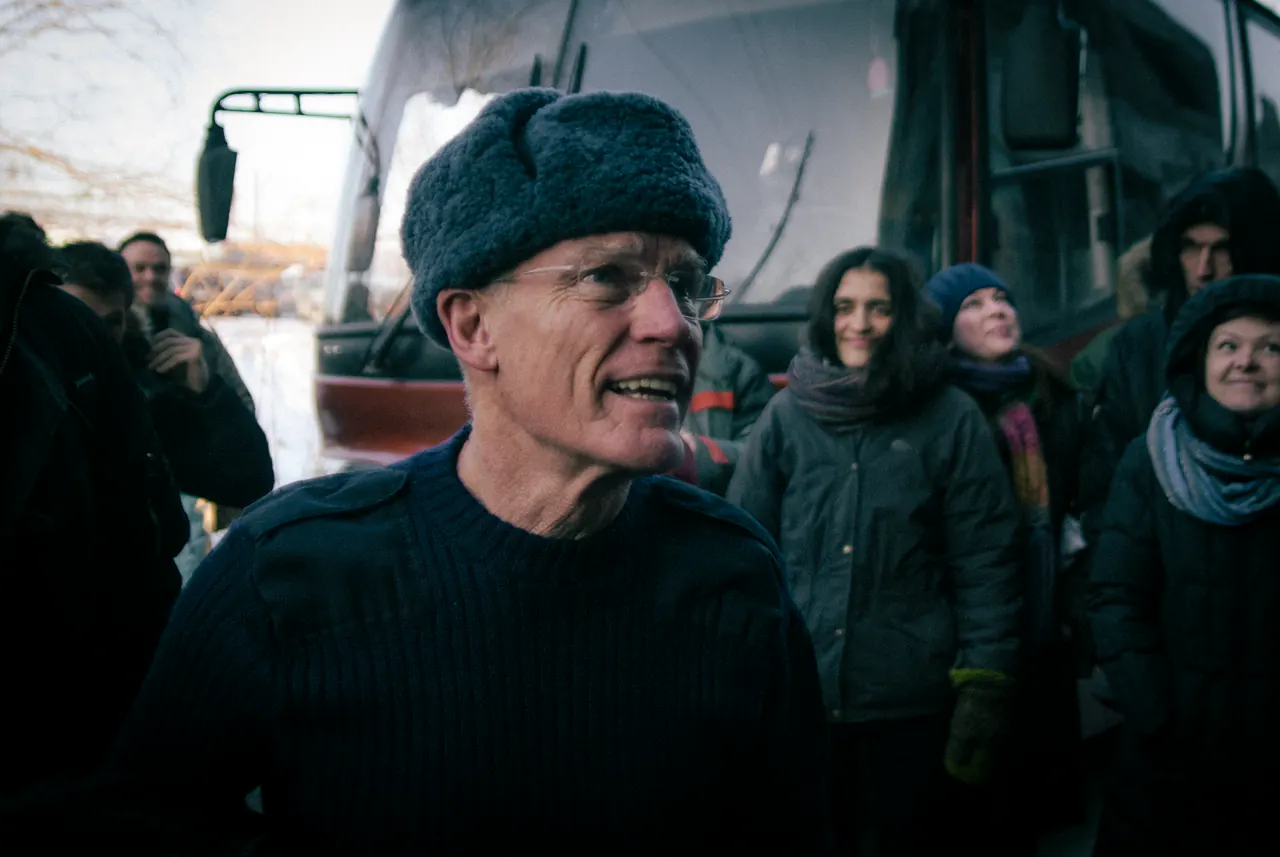 Lama Ole Nydahl in a fur hat smiles beside a minibus while travellers wait in the cold