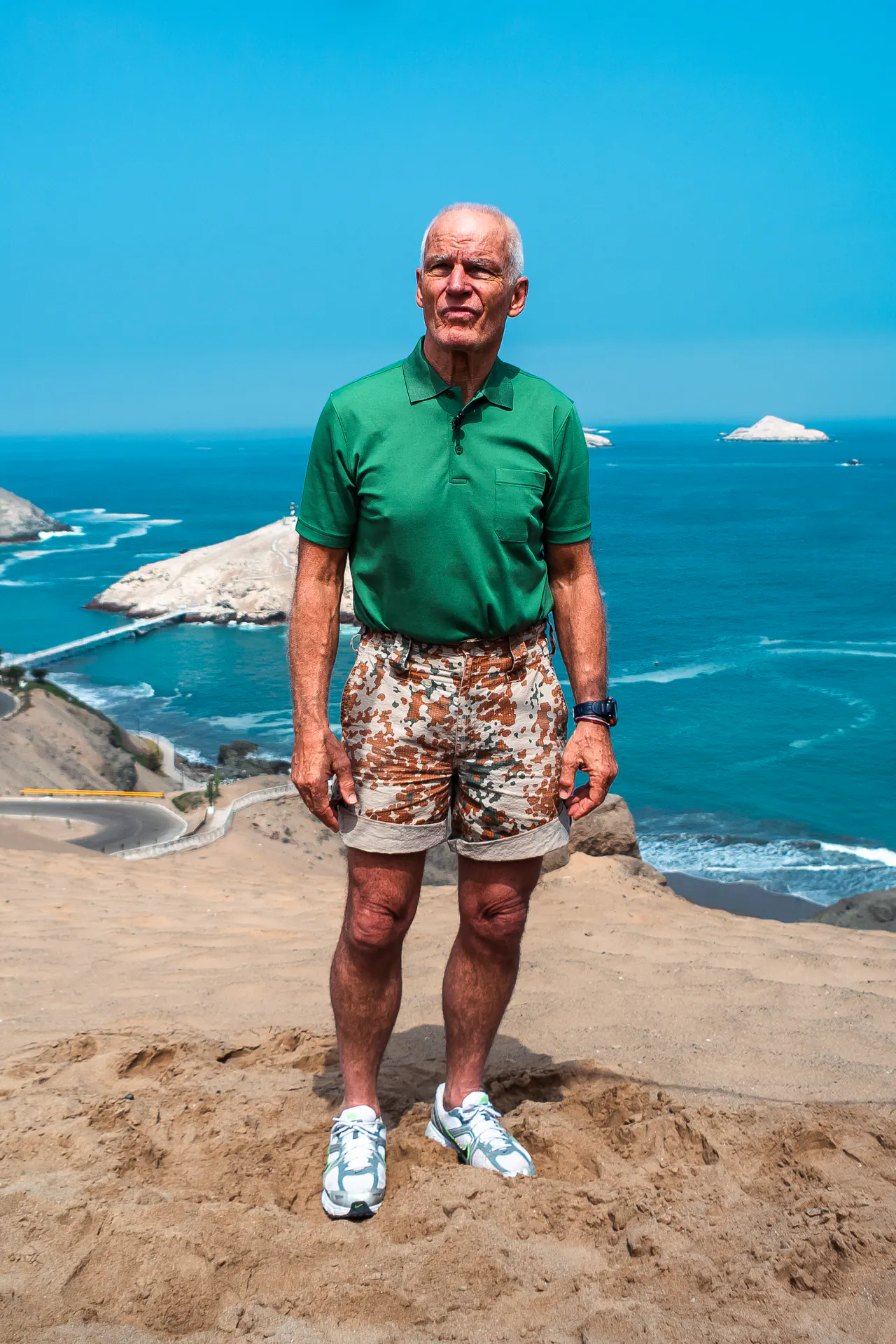 Lama Ole Nydahl in a green polo shirt stands on a sandy clifftop above the sea