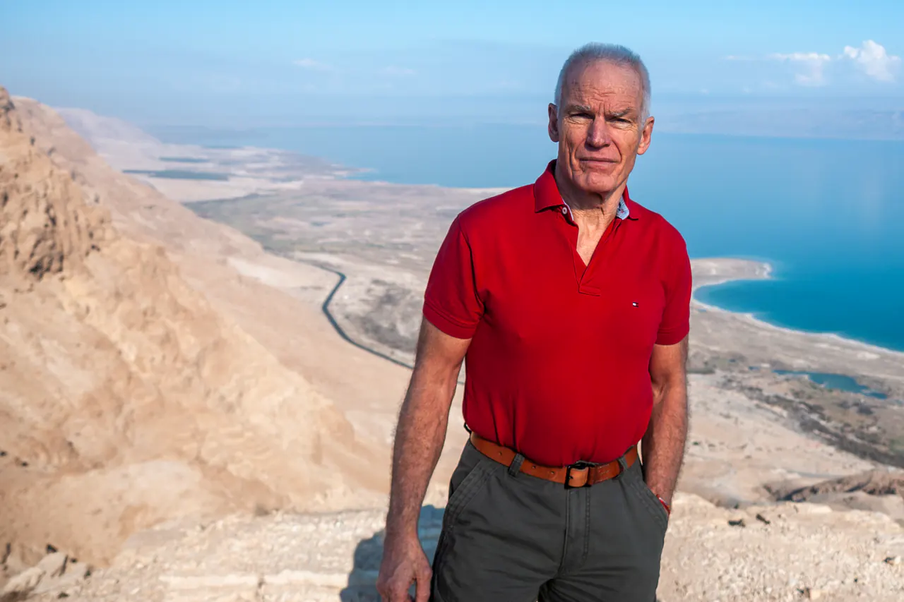 Lama Ole Nydahl in a red polo shirt stands on a ridge above a desert road and a distant sea