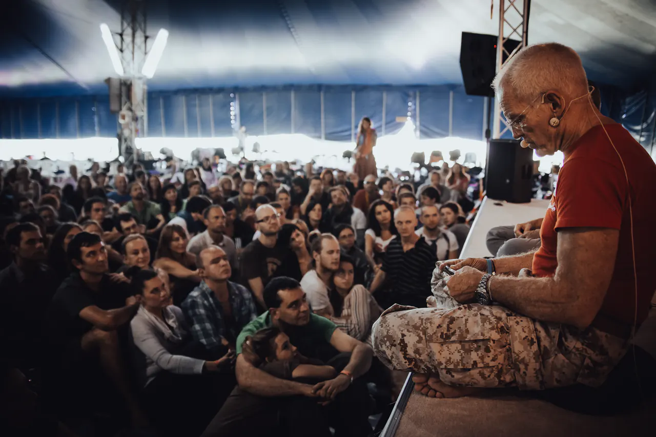 Lama Ole Nydahl sits cross-legged on a stage in a large tent, lecturing to a crowd seated on the floor