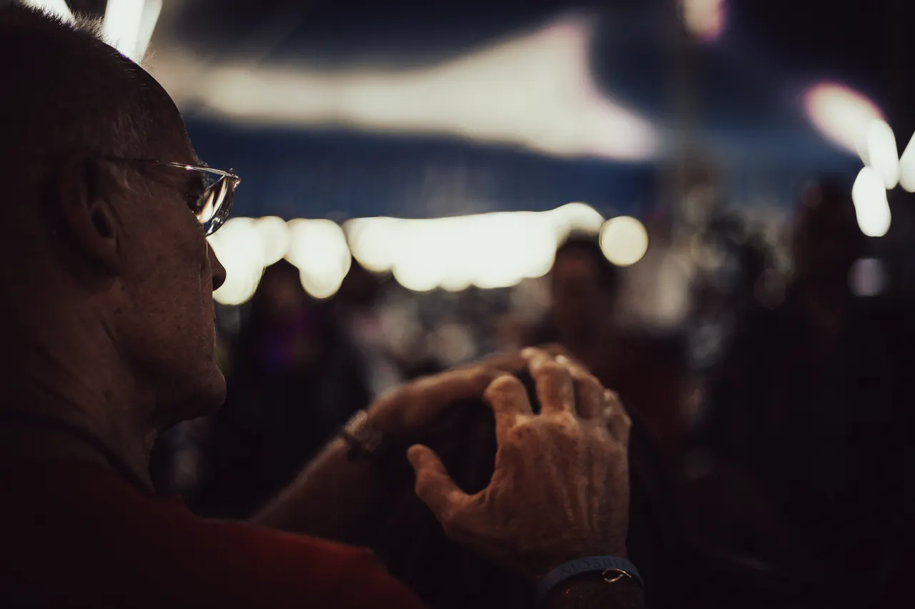 In near darkness, Lama Ole Nydahl sits with his hands folded, lights blurred behind him