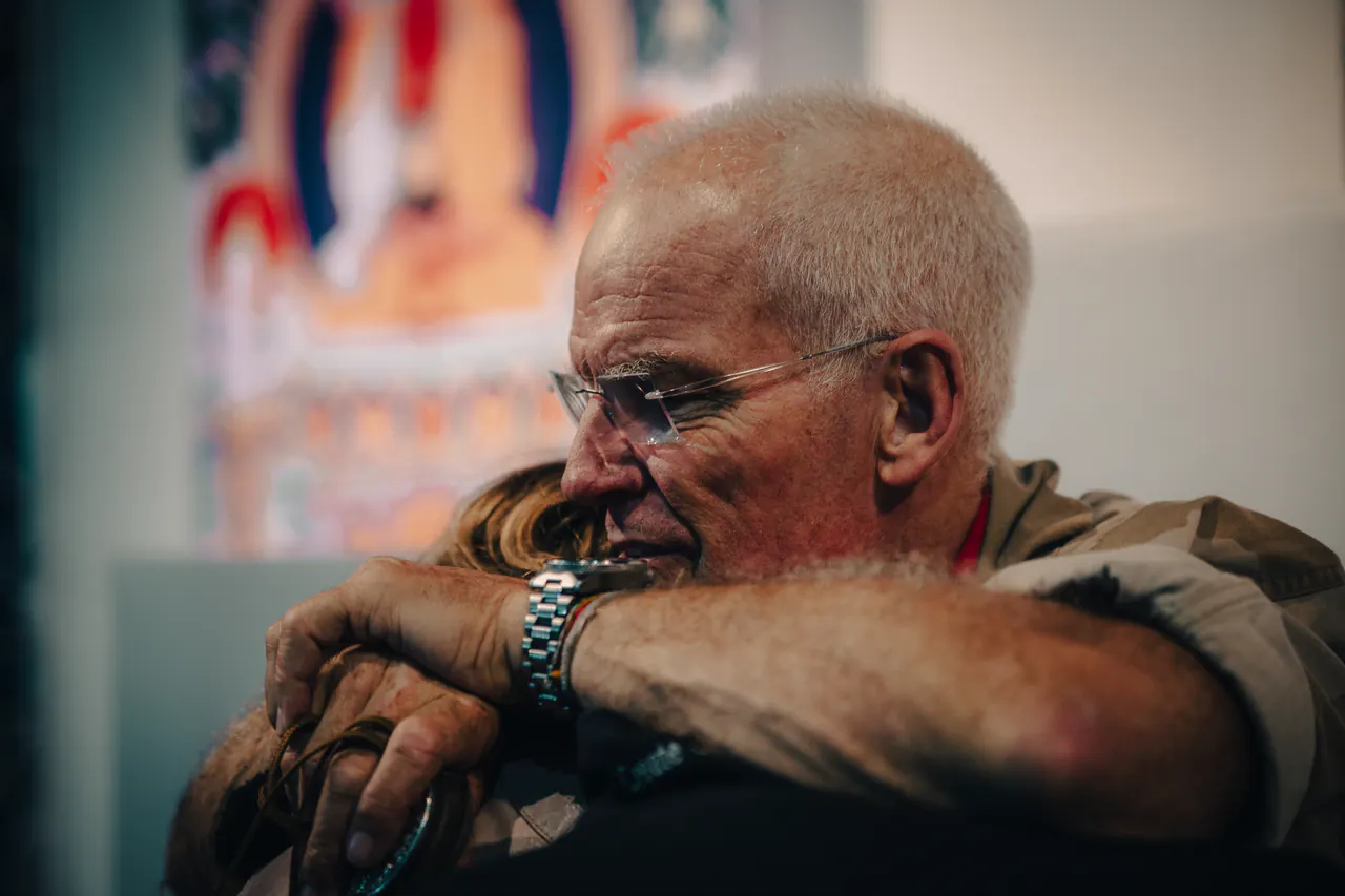 Lama Ole Nydahl rests his chin on folded arms, a thangka on the wall behind him