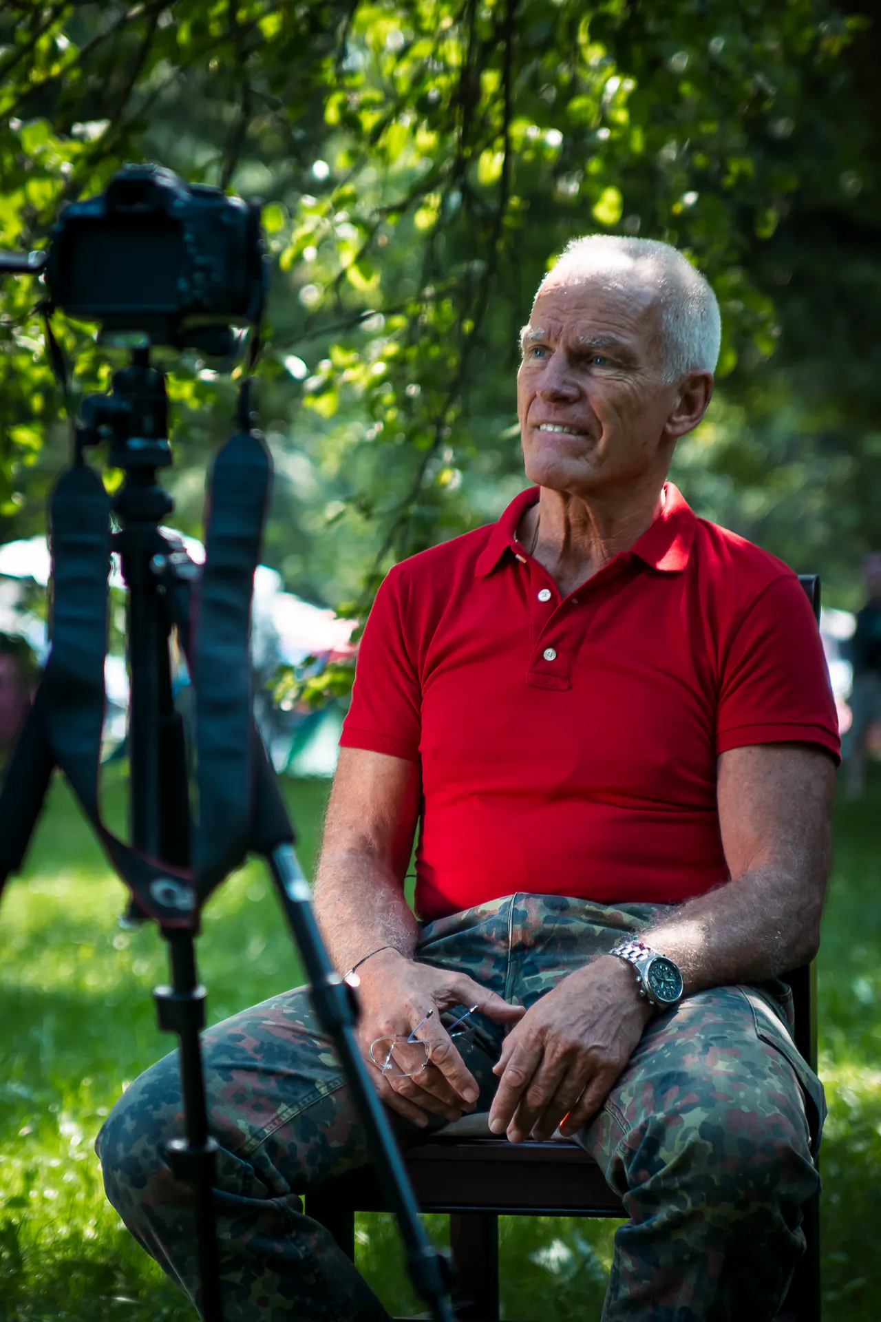 Lama Ole Nydahl sits on a chair under trees while a camera on a tripod records him