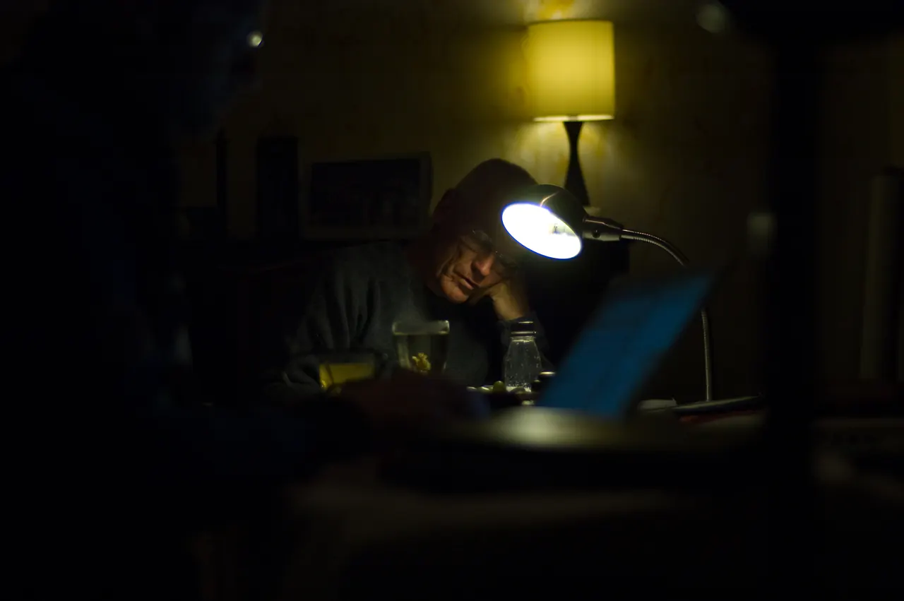Lama Ole Nydahl sits reading late at night, his face lit by a desk lamp, a laptop glowing in the dark foreground